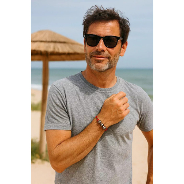 Homme d’une quarantaine d’années en t-shirt gris portant le bracelet rouge Gambier à trois perles de Tahiti, en bord de mer.